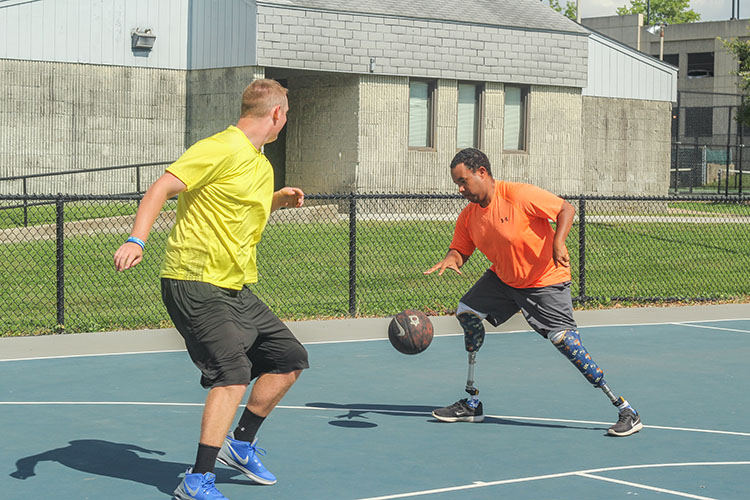 Two men are playing in an outdoor basketball court, both in brightly colored shirts. The man with the ball also has two prosthetic legs