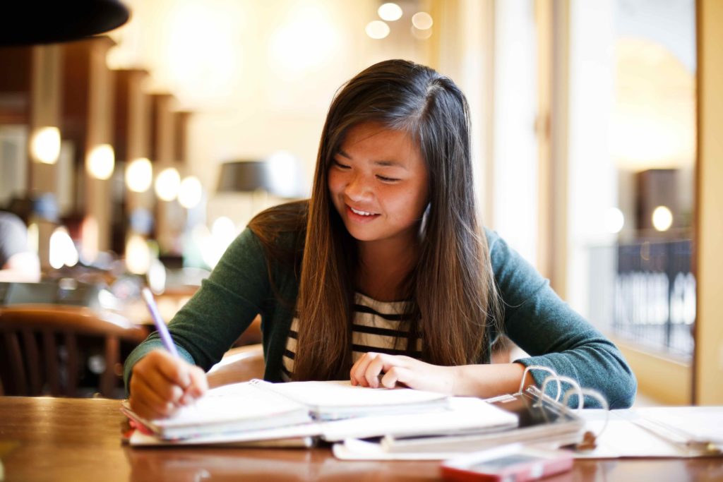 A student in the library writes in a notebook. She has long dark hair, a green sweater, and stripped tee.