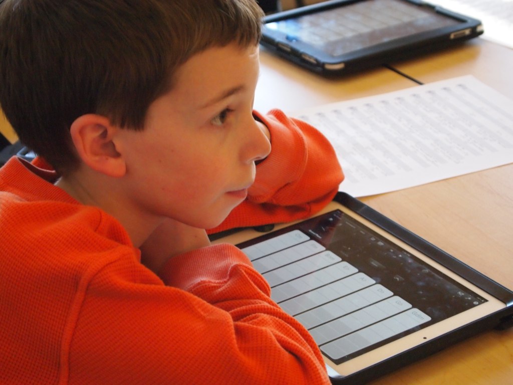 A student has an iPad in front of him, an AAC device. He is looking toward the front of the classroom. His hair is brown and his long sleeve shirt is orange.
